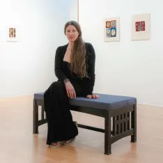 An art student sitting on a bench in a gallery. The exibit around her features her artwork.